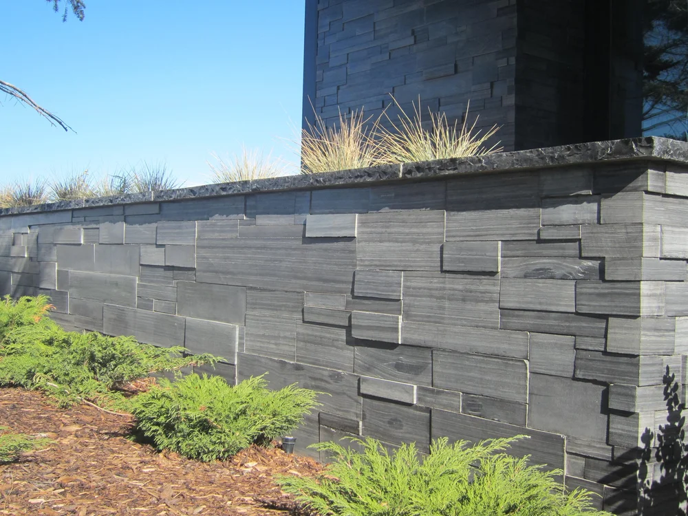 Black Rundle Stone builds a low capped garden wall in dark stacked courses above juniper and ornamental grass.