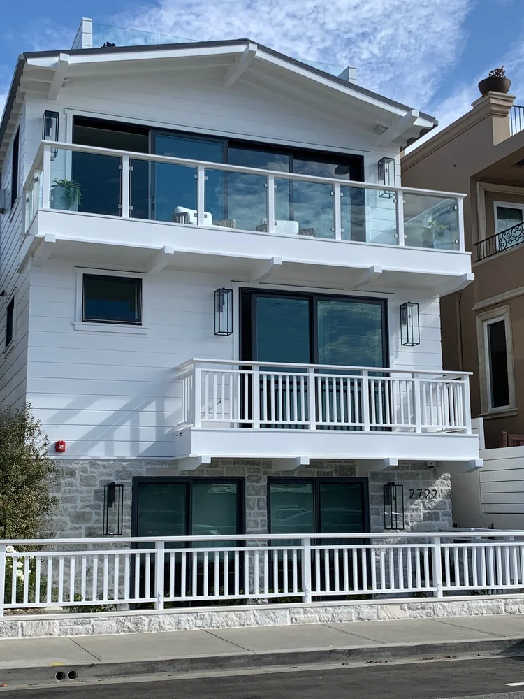Blue grounds a three storey beach house, the stone base reading against white siding and glass rails above.
