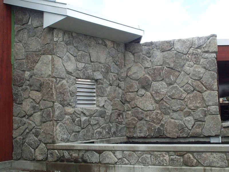 Larger Grizzly mosaic pieces clad a low exterior wall with a louvered vent, a water channel below.