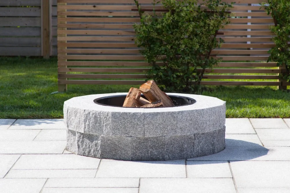 A round Woodbury Gray fire pit holds split logs on a flamed paver terrace.