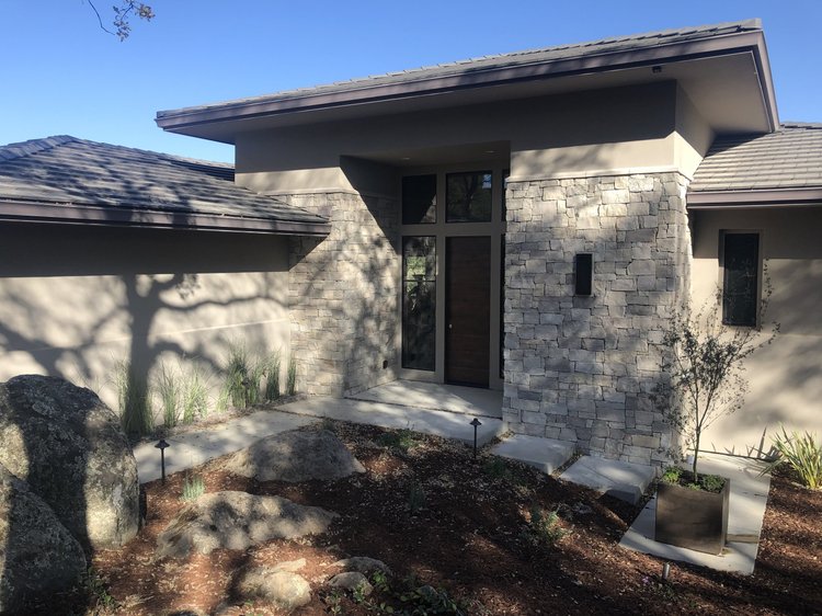 Mayville wraps the entry mass of a low-slope house, boulders and grasses banked below.