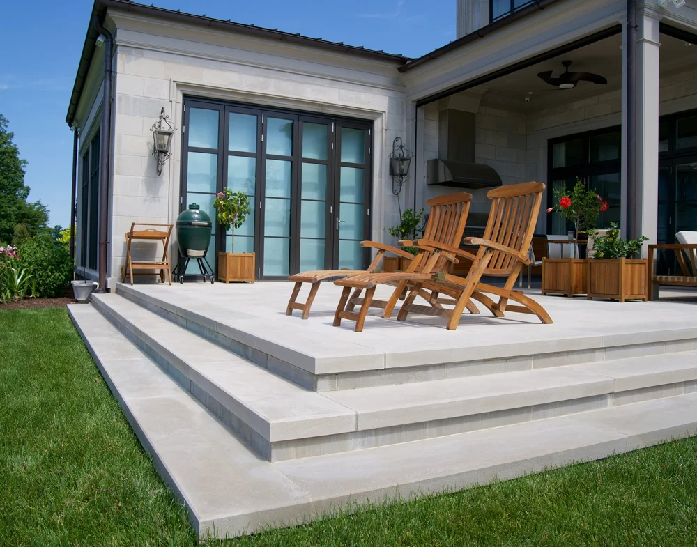Indiana Limestone Gray paving and two broad step courses lift a terrace above the lawn, teak loungers set out under the eave.