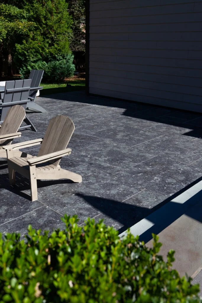 Flamed Saint Henry Black paving running to the corner of a dark clapboard wall, Adirondack chairs at the edge and a clipped hedge in the foreground.