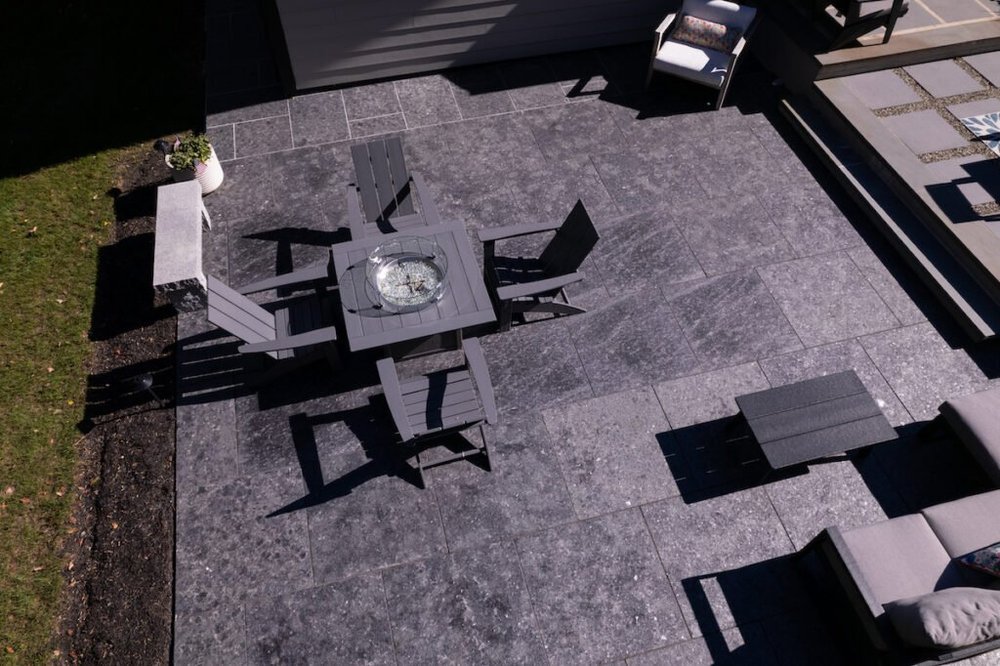 Overhead view of a back terrace paved in flamed Saint Henry Black, a fire table and Adirondack chairs at its center with lawn along one edge.