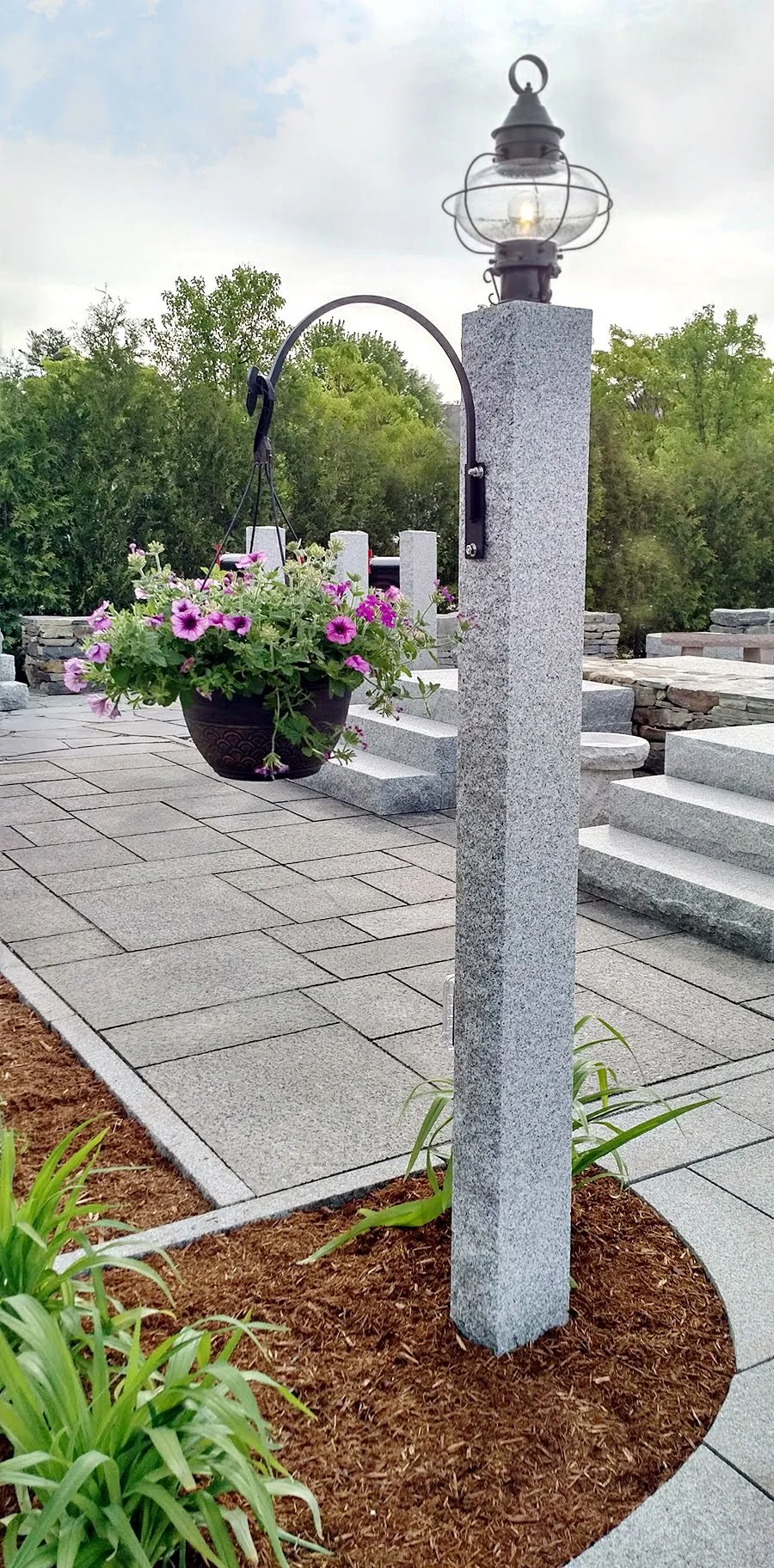 A square Woodbury Gray light post carries a lantern and a hanging basket over paving.