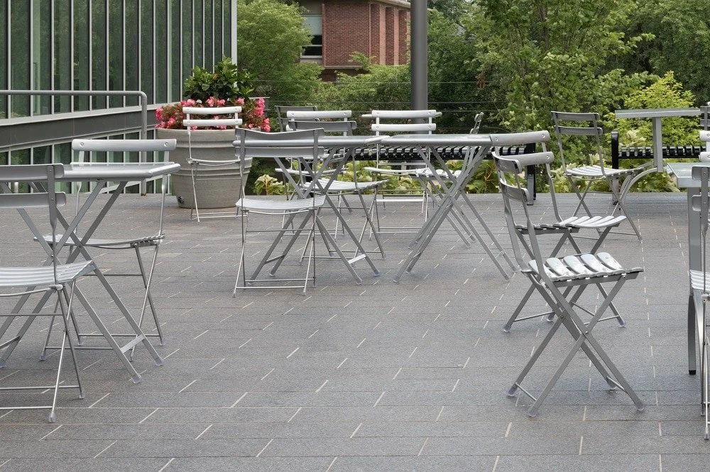 Cambrian Black paving carries a cafe terrace of folding bistro tables and chairs below a planted railing.