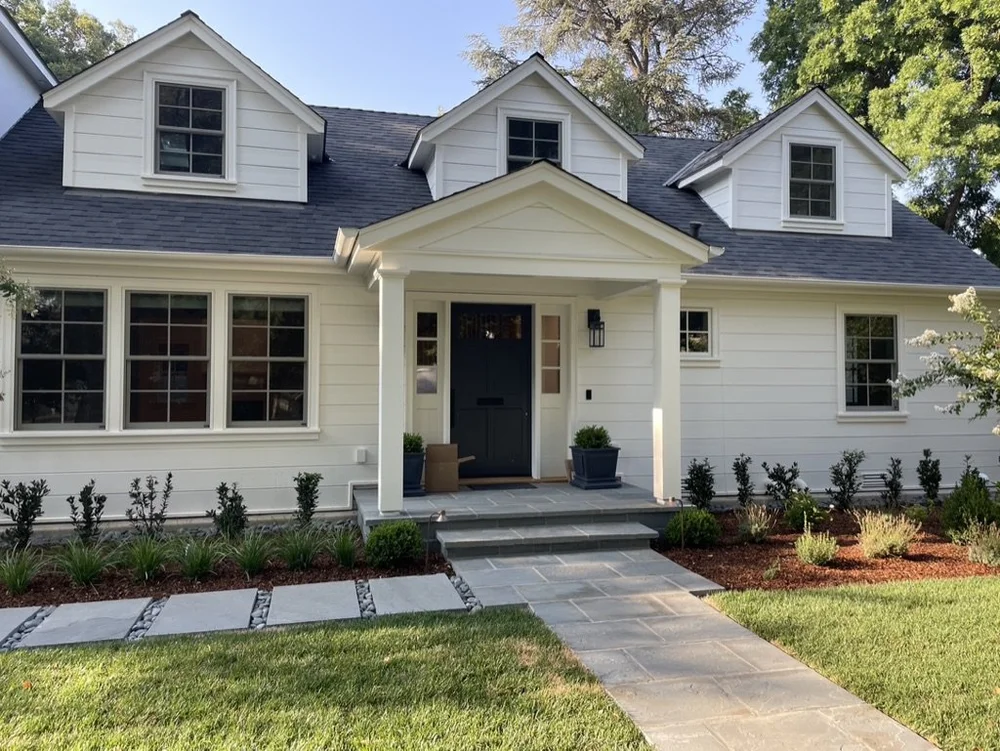 Bluestone paves the front walk and entry steps of a white dormered house, stepping stones carrying on across the lawn.