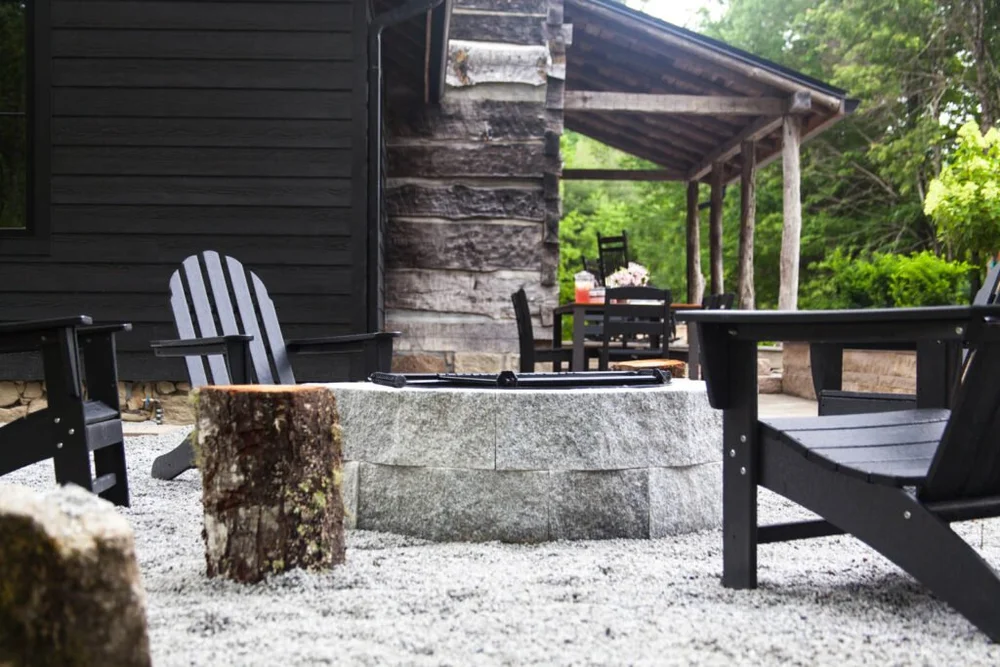 A rough faced Woodbury Gray fire pit sits on white gravel before a dark cabin and its stone chimney.
