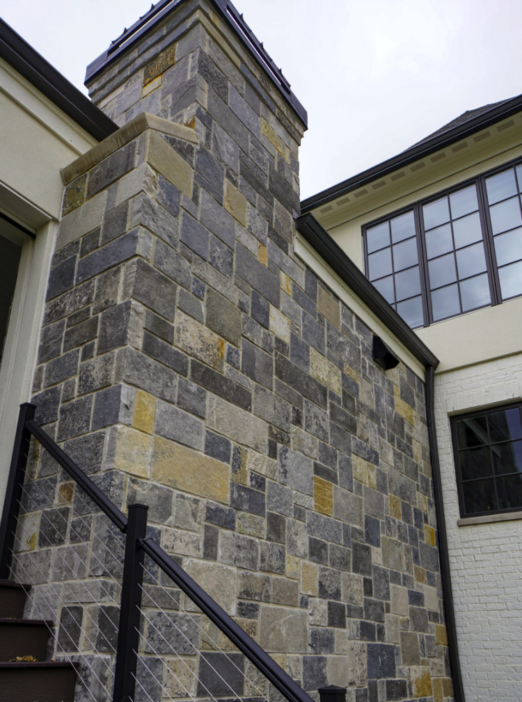 Graphite castle veneer builds a capped chimney mass against white siding, a black stair rail crossing below.