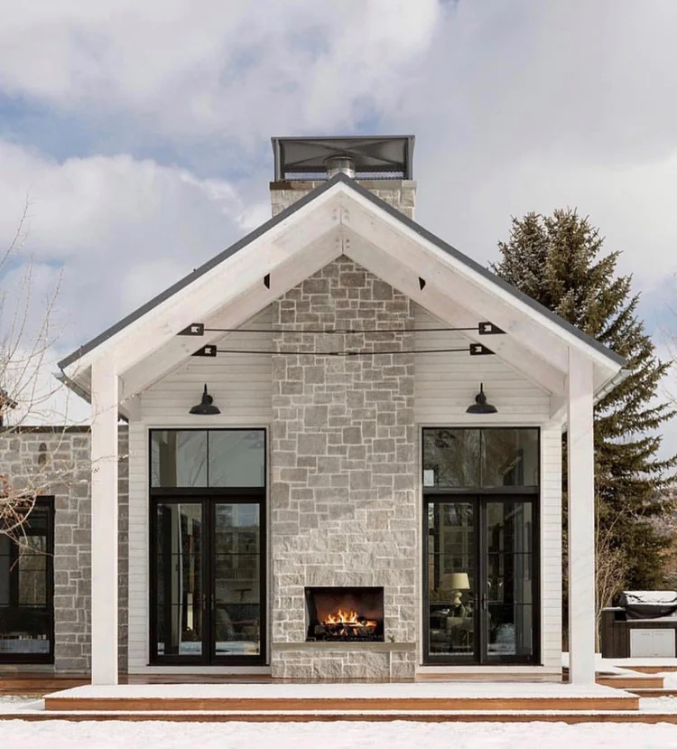 Flint Hills Gray builds the chimney mass of a gabled outdoor room, its firebox lit between black framed doors.