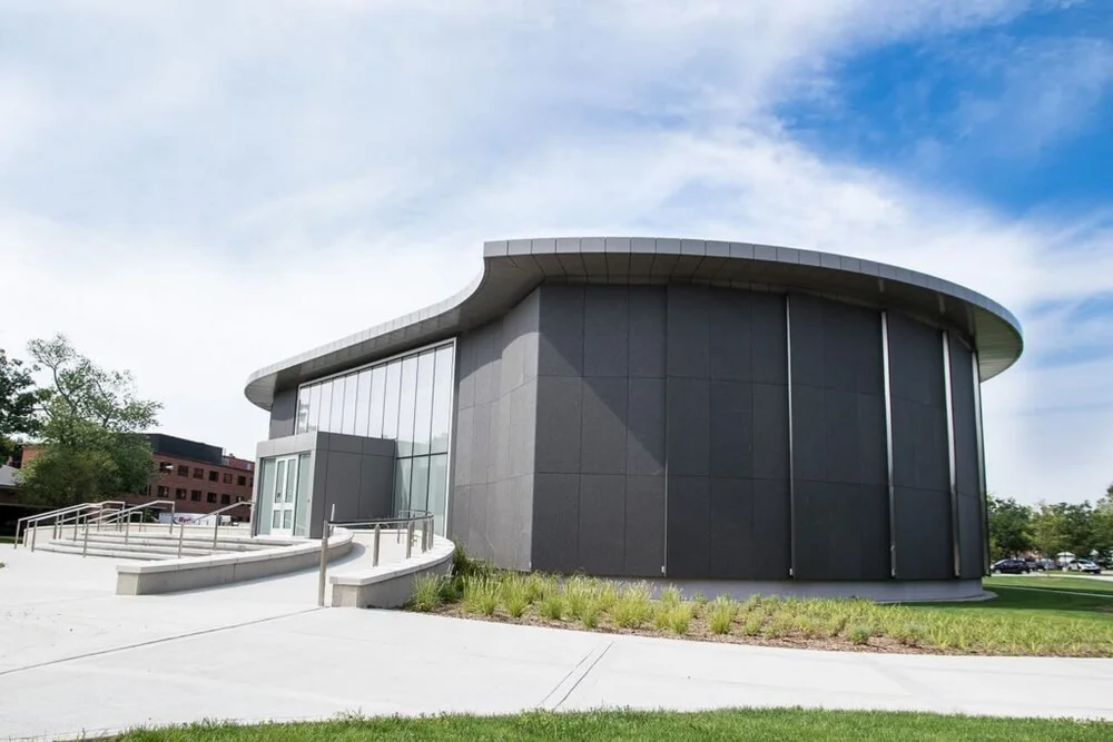 Cambrian Black in flamed panels wraps the curved drum of a museum, meeting the glazed entry at the ramp.