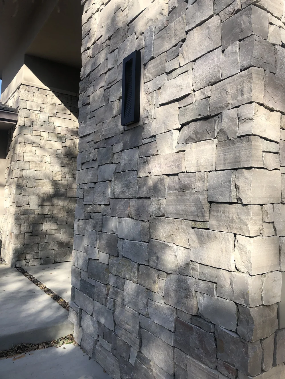 A Mayville clad entry column rises beside a slim sconce, its dry-stacked courses catching hard afternoon shadow.