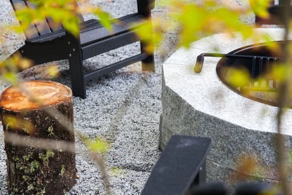 A solid granite fire bowl and its grate seen through autumn leaves, black chairs drawn up on gravel.
