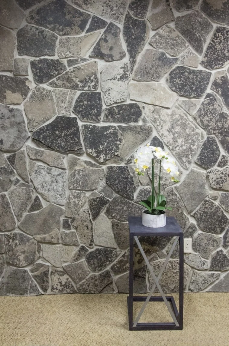 Graphite mosaic covers an interior wall in large angular pieces behind a slim plant stand.