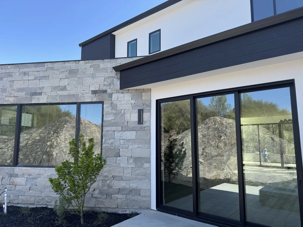 Charcoal coursed veneer in split face runs the ground floor between black-framed windows and sliding doors.