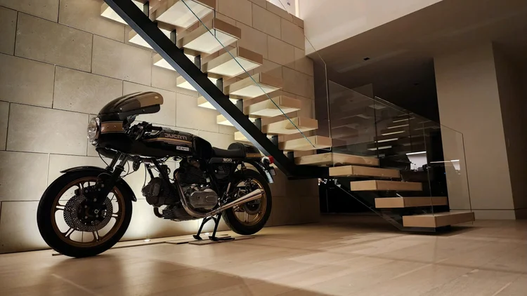 Light from the floating treads rakes across an Indiana Limestone Buff panel wall, a motorcycle parked beneath the stair.