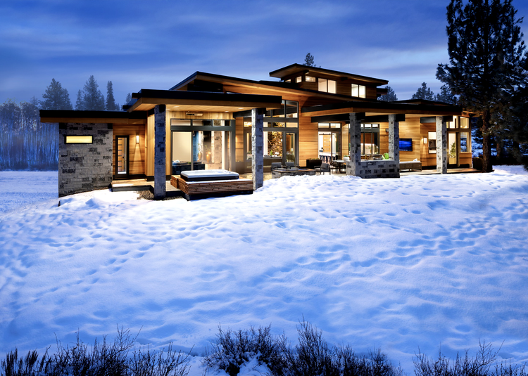 A Charcoal chimney mass and veneer piers ground the home at dusk across an open snowfield.