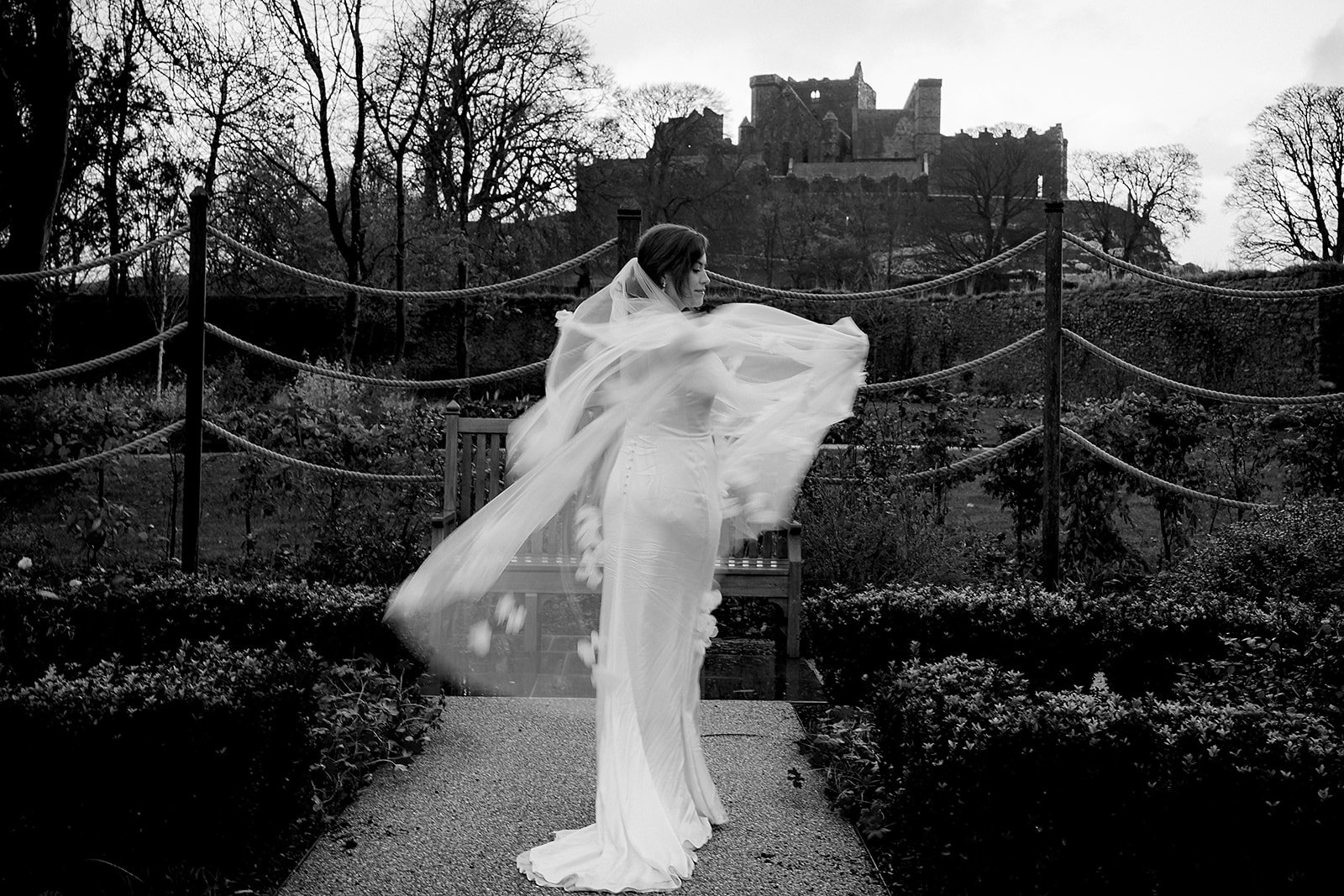 Black and white film portrait of a bride on a castle path, her veil lifting in the wind