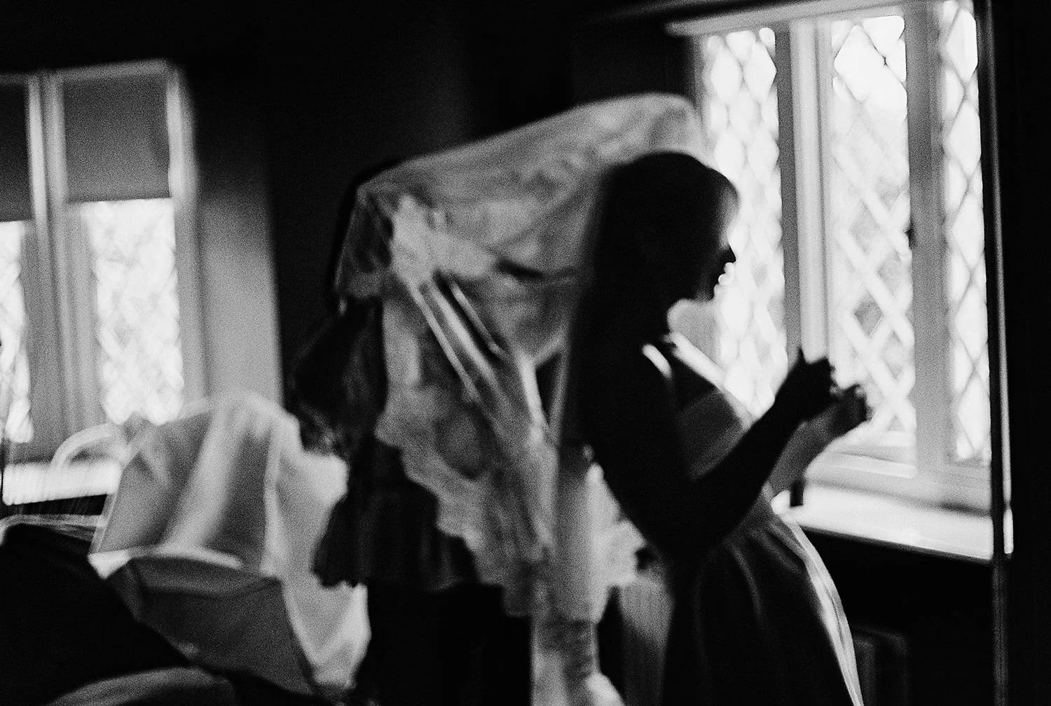 Black and white film photograph of a bride silhouetted at a leaded window as her veil is arranged