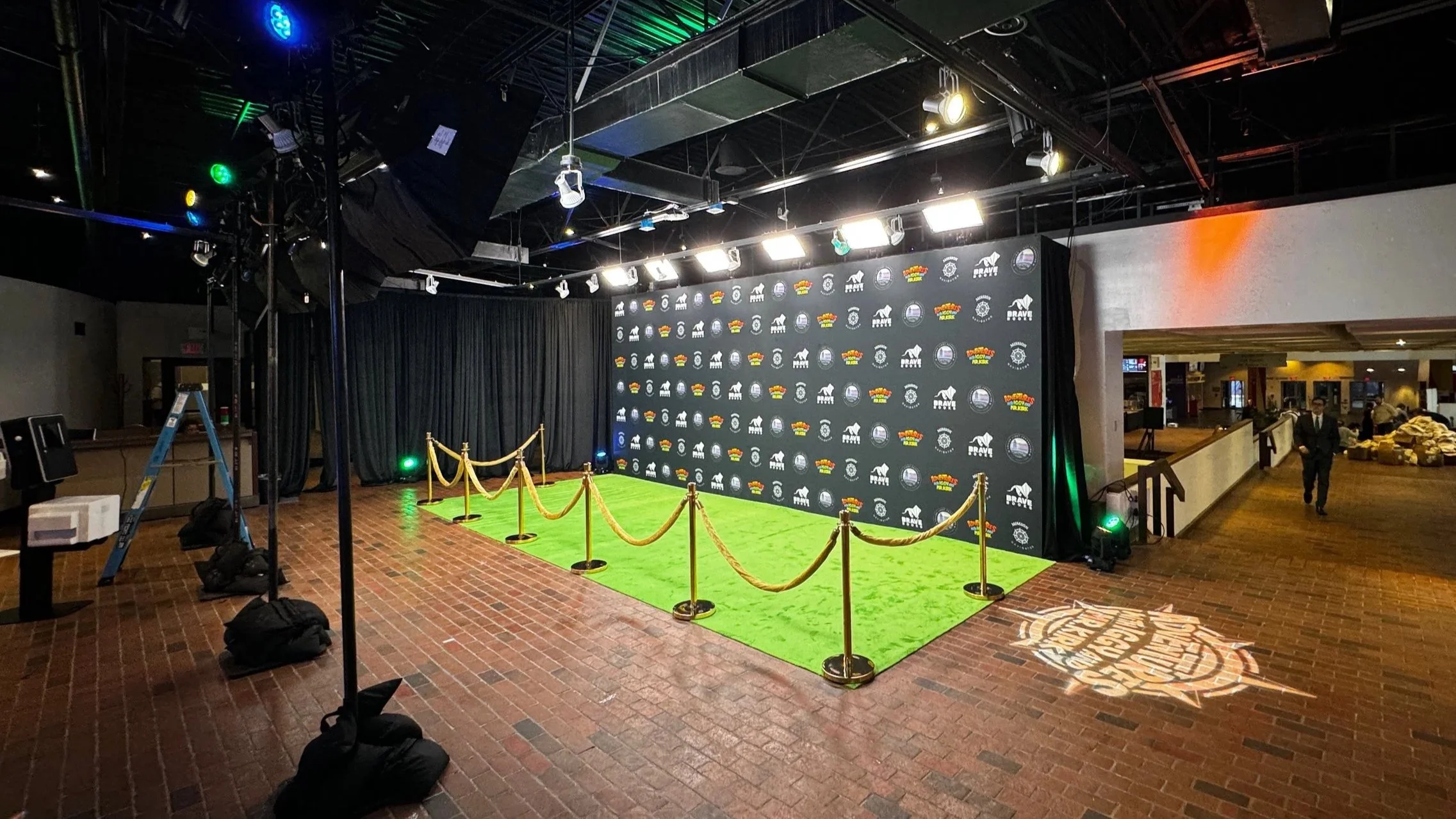 Step and repeat premiere setup by The Camera EYE — fully lit branded backdrop with green carpet and gold rope stanchions, overhead LED panel array providing even illumination, branded gobo projected on brick floor, green and orange uplighting flanking the backdrop