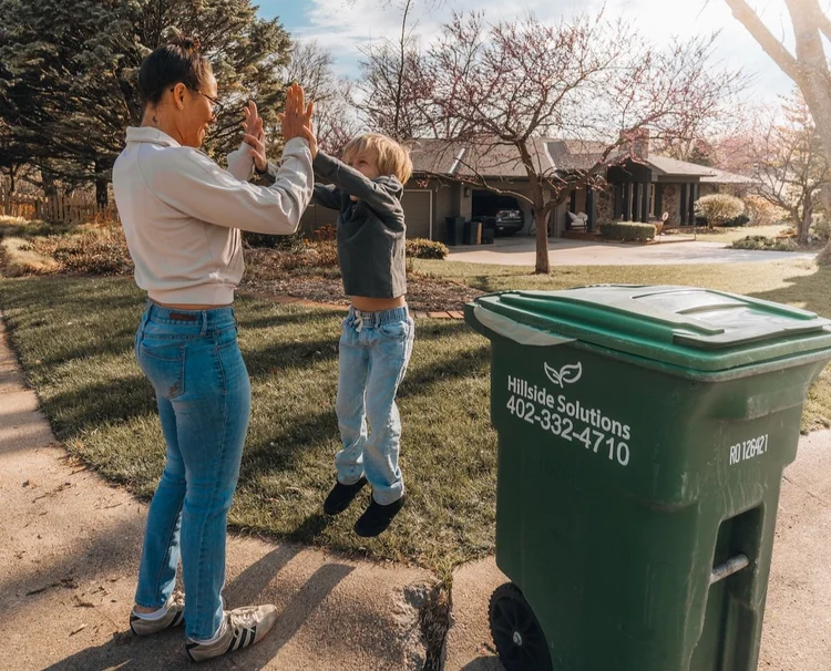 A mom and her kid high-five next to a Hillside Solutions Compost Club cart at the curb