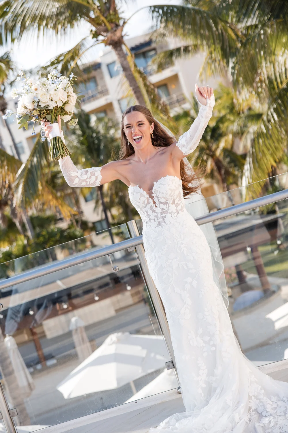 A bride laughing with her bouquet raised on a resort terrace framed by palms
