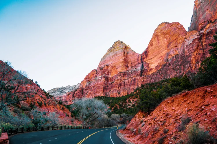 A paved road curves beneath layered red sandstone cliffs, with a reddish roadside slope, bare trees and green canyon vegetation.