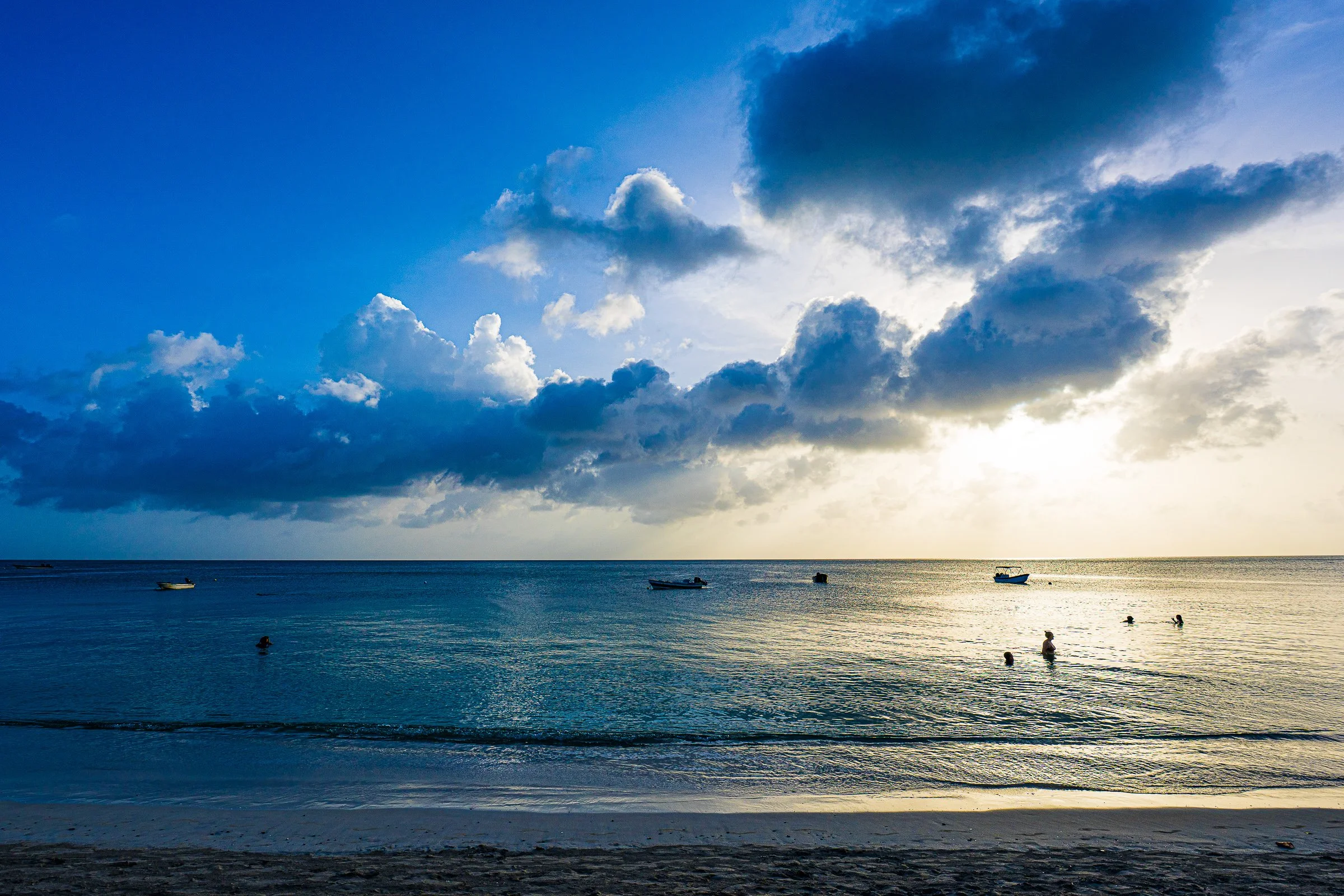 A tranquil Caribbean beach with distant swimmers under a dramatic blue cloudbank.
