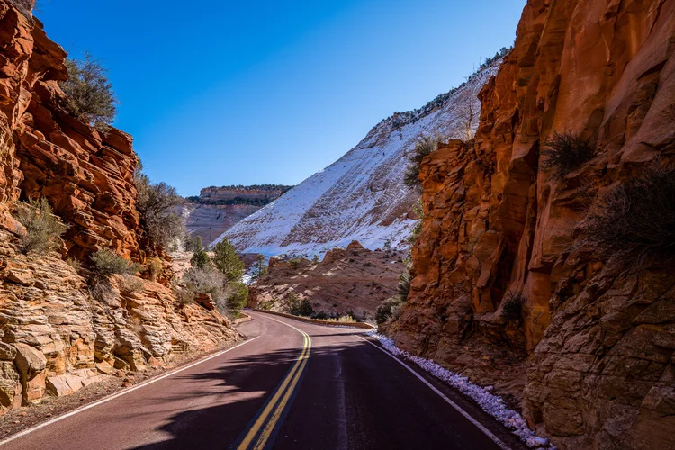 A two-lane road curves between orange sandstone walls, with a snow-covered slope in the distance under a clear blue sky.