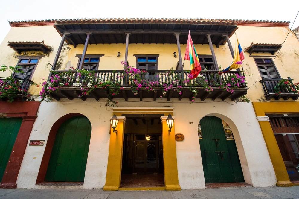 Warm lanterns frame a doorway beneath a flower-lined balcony and colorful flags.