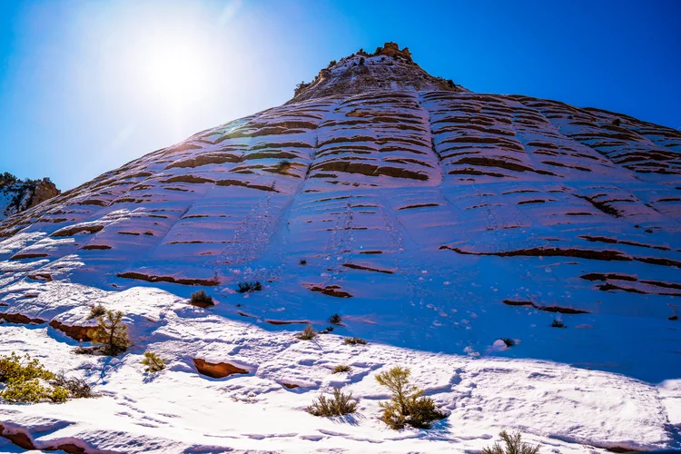Sunlight breaks over a snow-covered sandstone slope, with exposed red rock ledges and low shrubs beneath a deep blue sky.