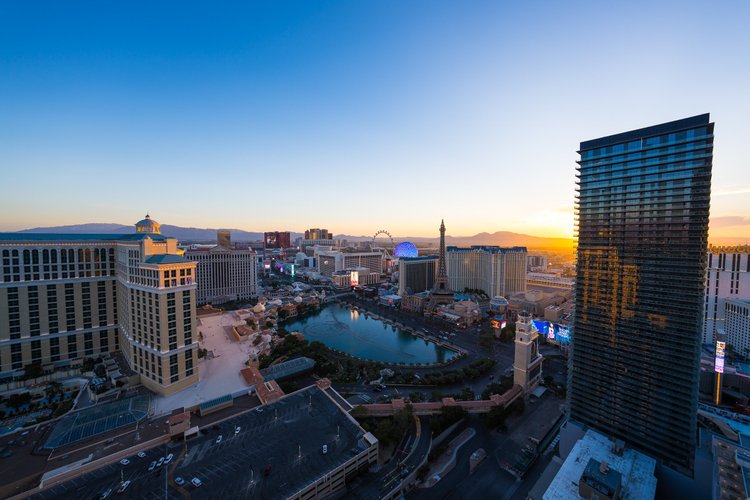 Elevated view of the Las Vegas Strip and a turquoise lake beneath a pale sunset sky, with a dark glass tower to the right.
