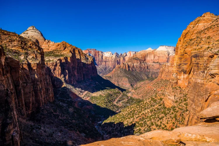 A sweeping canyon view from a sandstone overlook, with towering red and cream cliffs, a green valley and a winding road beneath a blue sky.