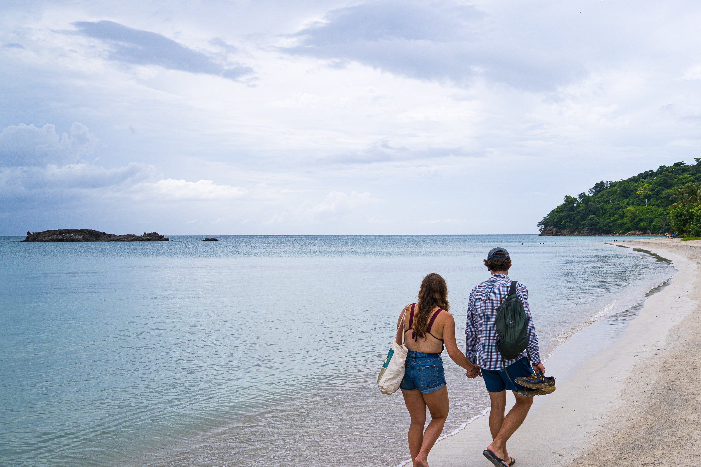 Two travelers walk hand in hand along pale sand beside calm shallow water.