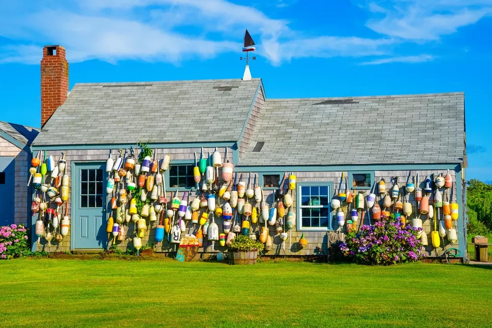 Weathered shingled Nantucket cottage adorned with many colorful fishing buoys under summer blue sky.
