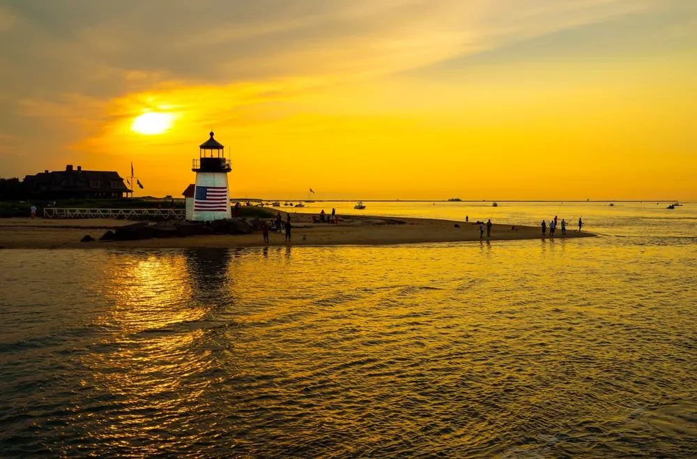 At golden sunset, Brant Point Lighthouse rises beside a sandy Nantucket shoreline with scattered beachgoers.