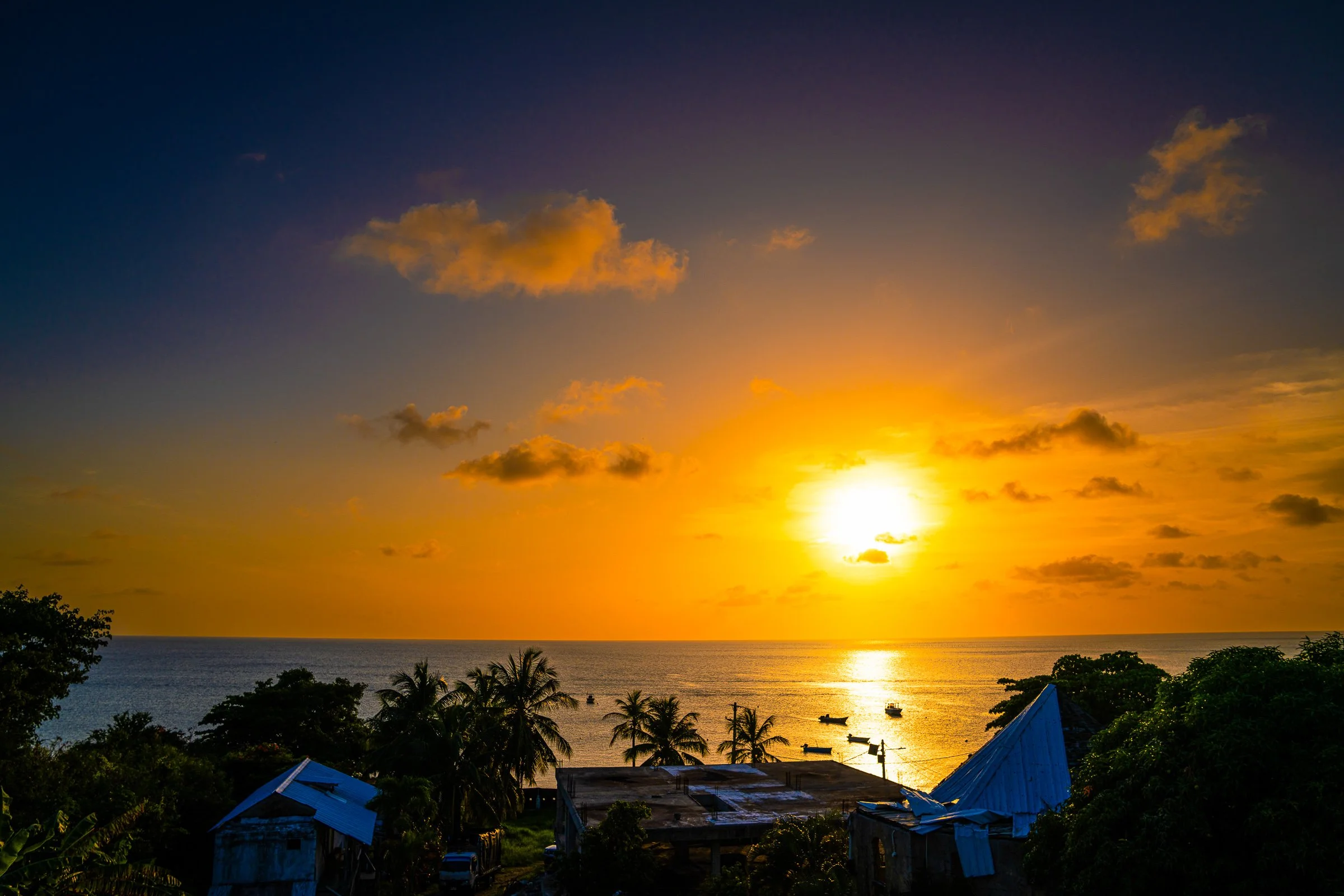 Orange-gold sunset beyond island rooftops, palms and small boats in the bay.