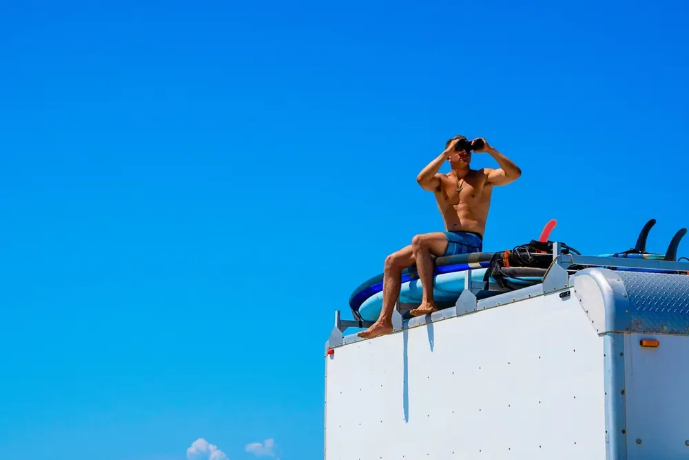 A person with binoculars sits above a vehicle carrying surfboards against a saturated blue sky.