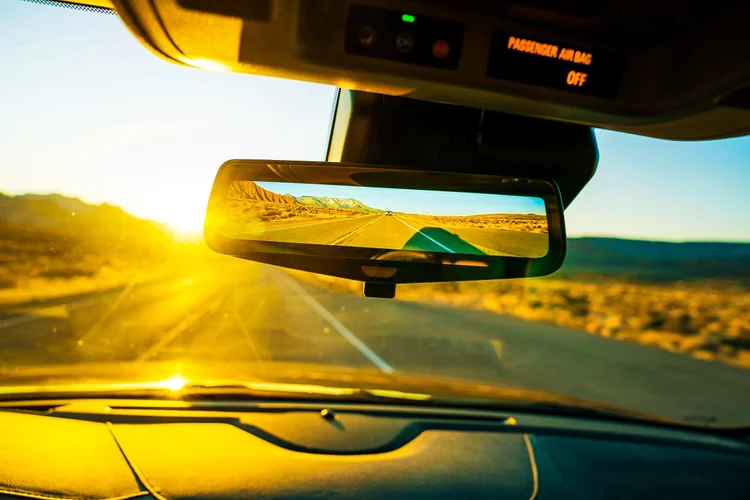 Golden sunlight fills a view through a car windshield, while the rearview mirror reflects an open desert road and the dashboard fills the foreground.