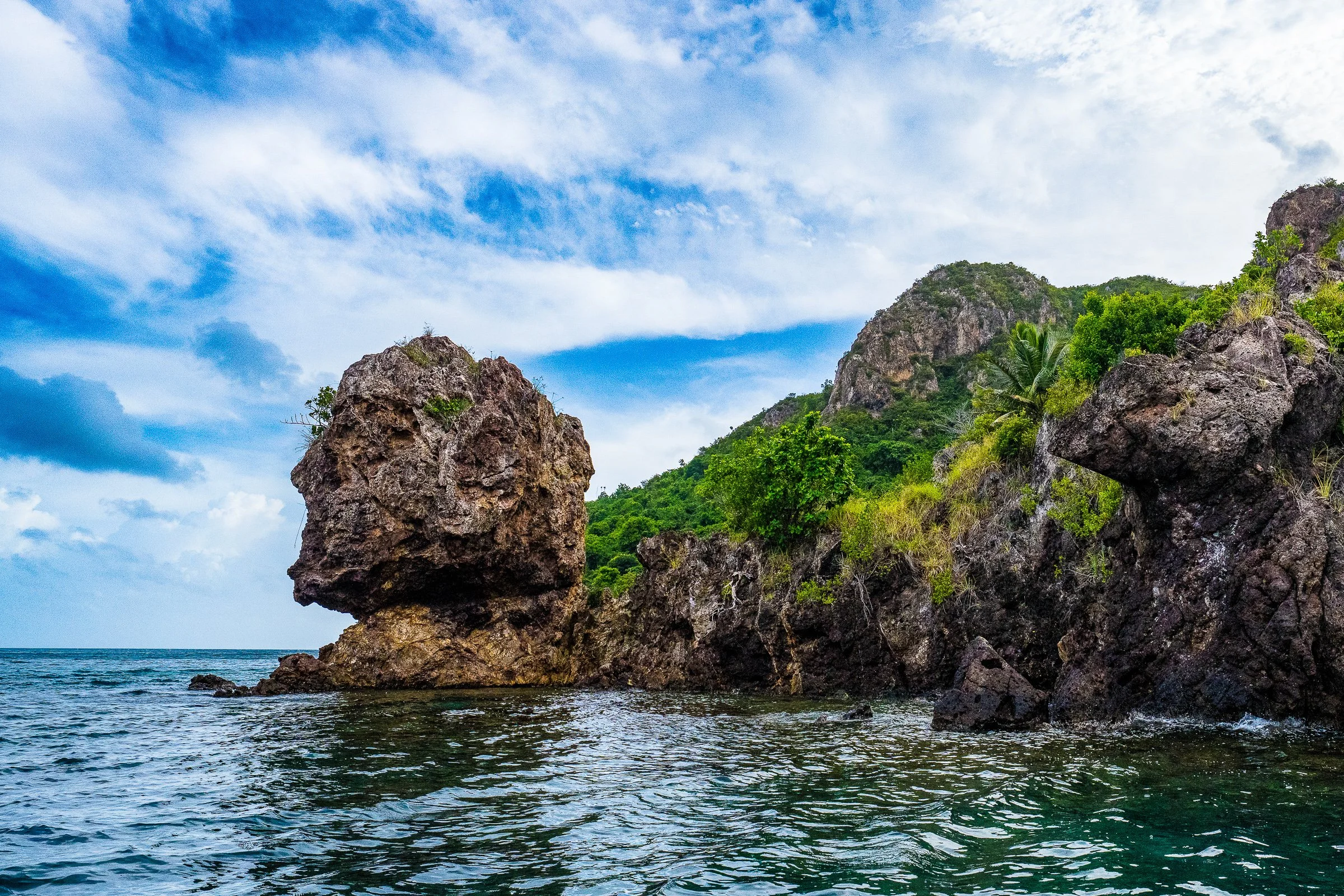 Sculptural weathered coastal rocks rise against green hills and a dramatic cloud-filled sky.