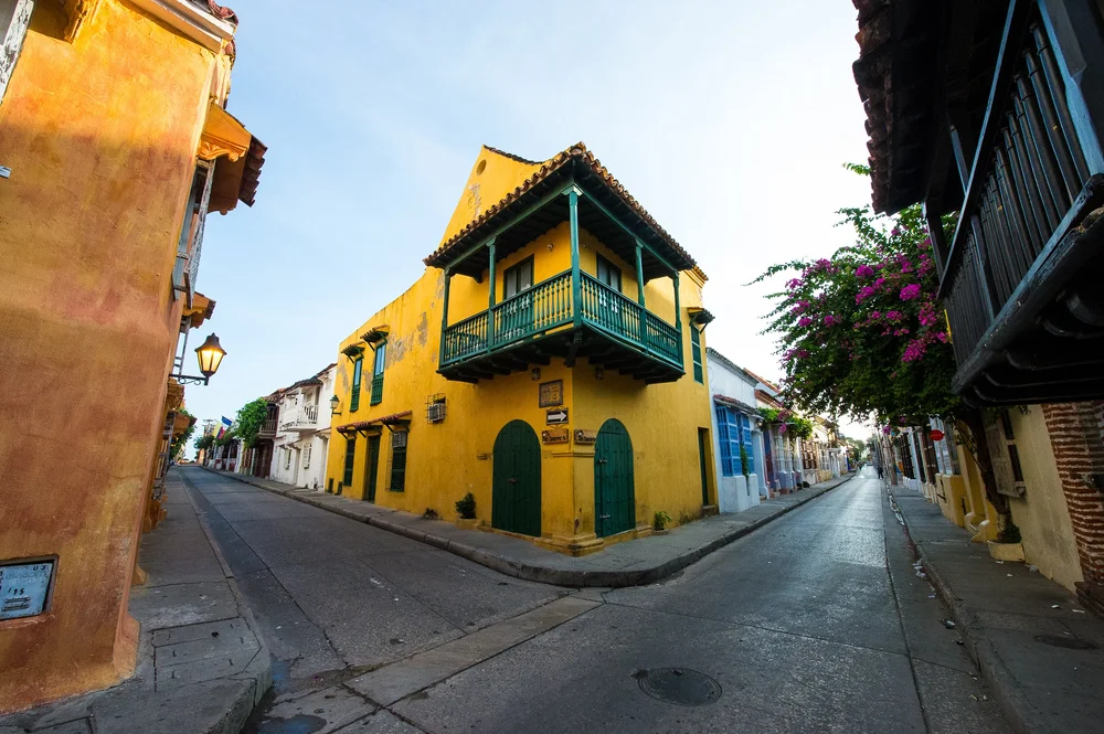 Green doors and a green wooden balcony accent a sunlit yellow corner house.