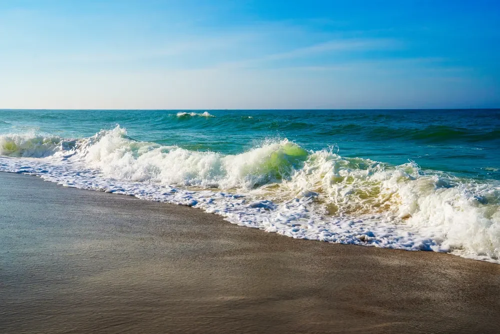 White Atlantic surf crashes diagonally onto a sandy Nantucket beach beneath blue skies.