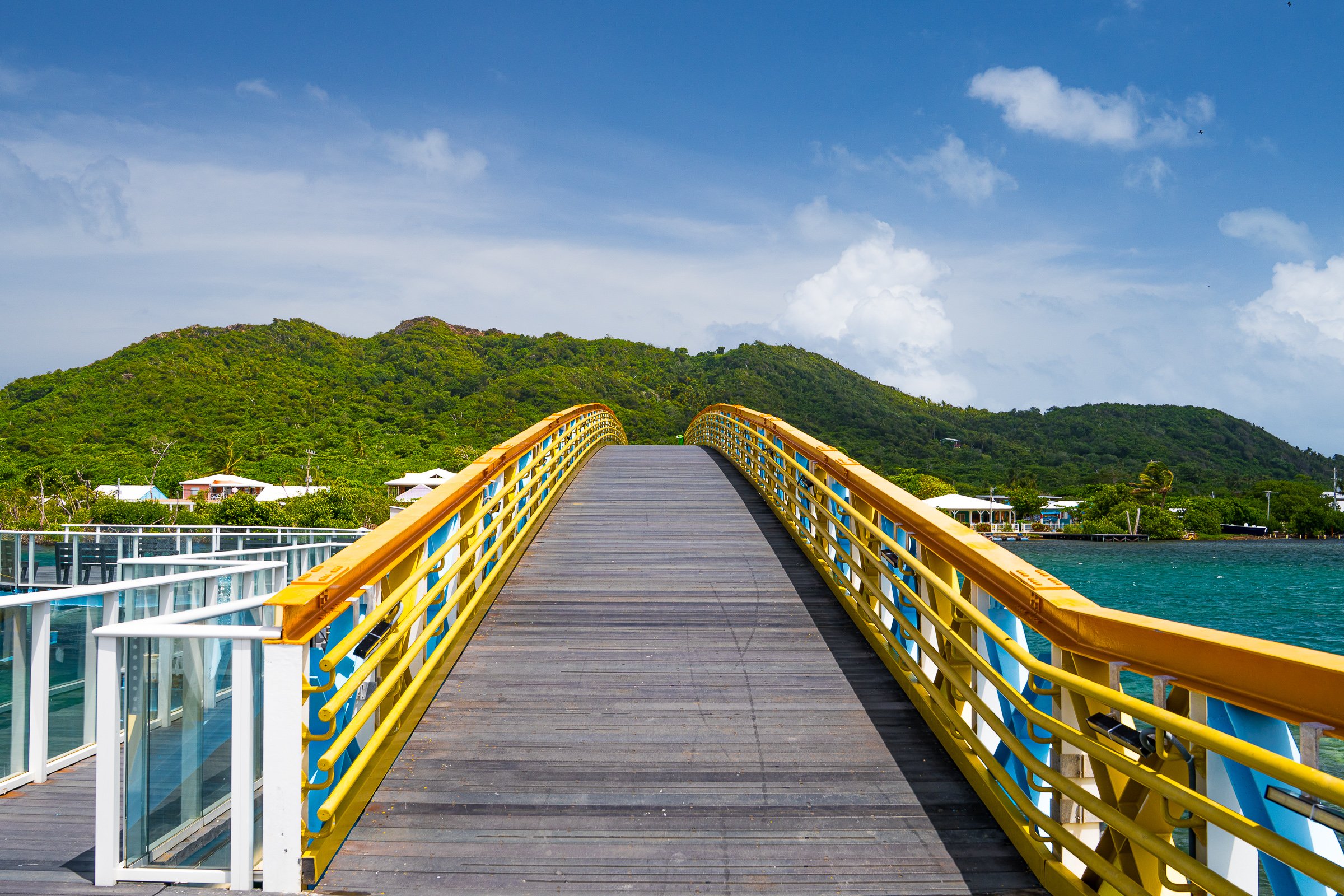 A yellow-railed wooden bridge crosses brilliant blue island waters toward green hills.