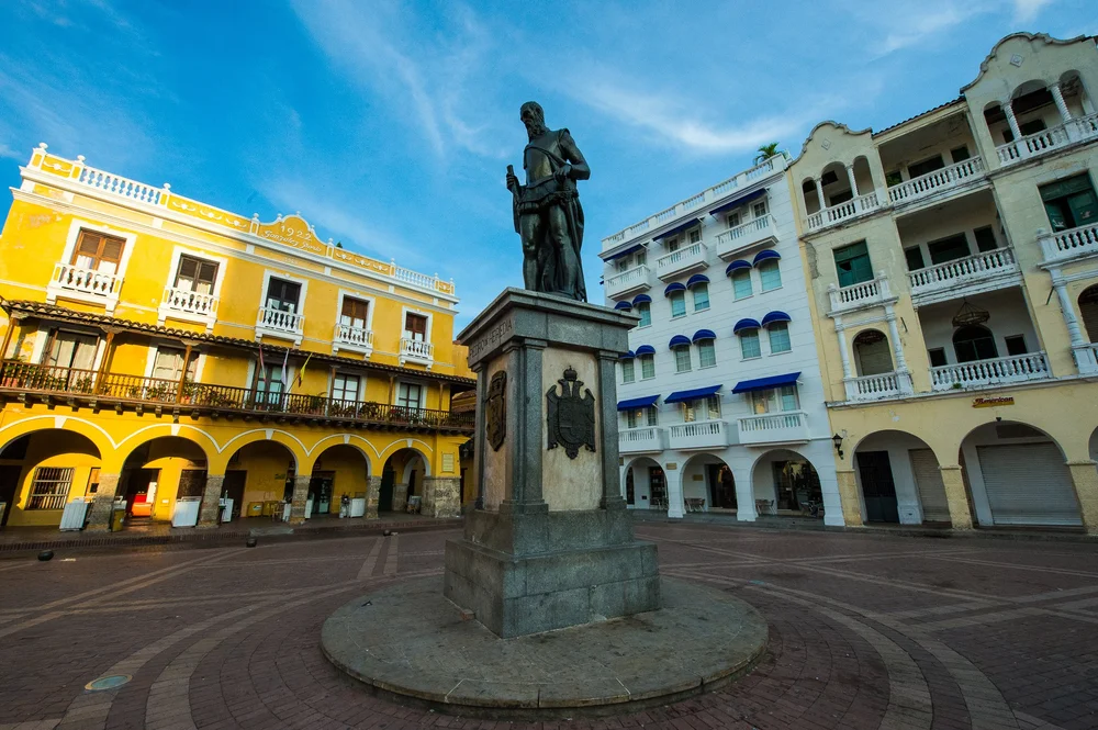 A statue stands in a historic square surrounded by colorful colonial buildings and open sky.
