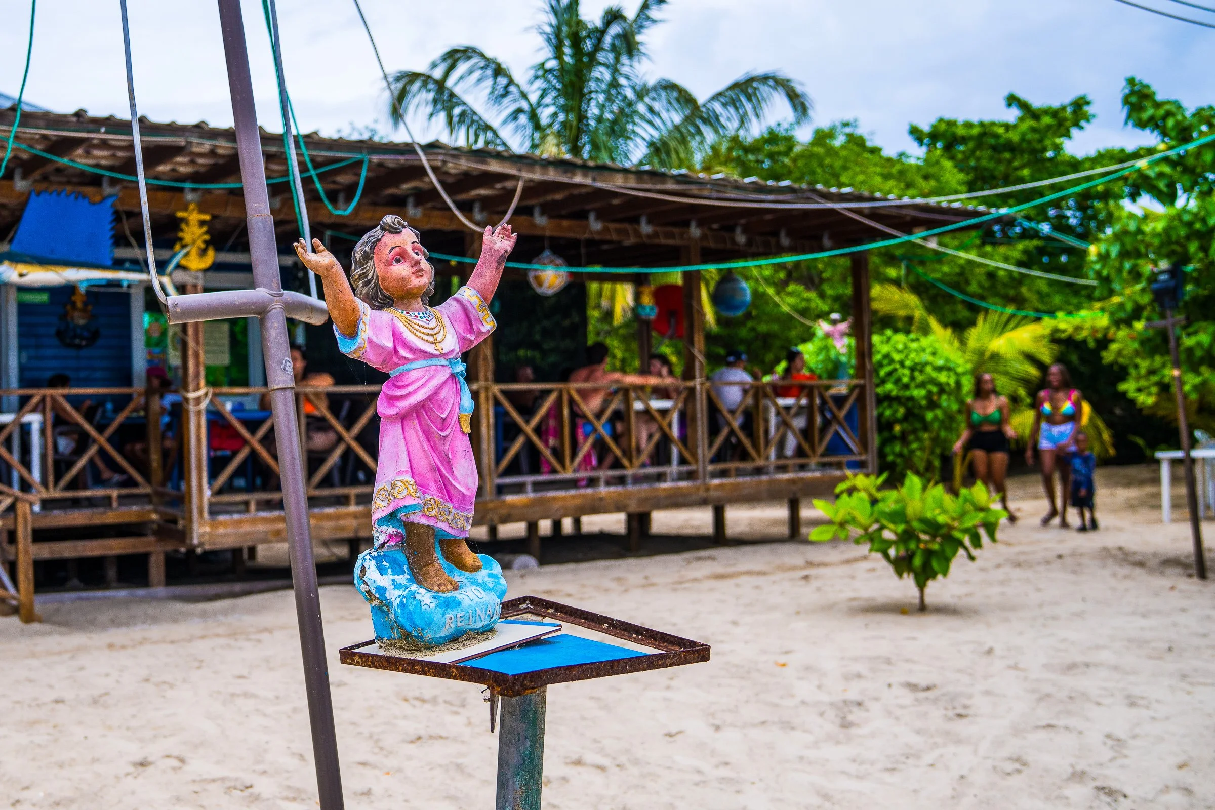 A small painted religious figure stands in front of a rustic open-air gathering place.