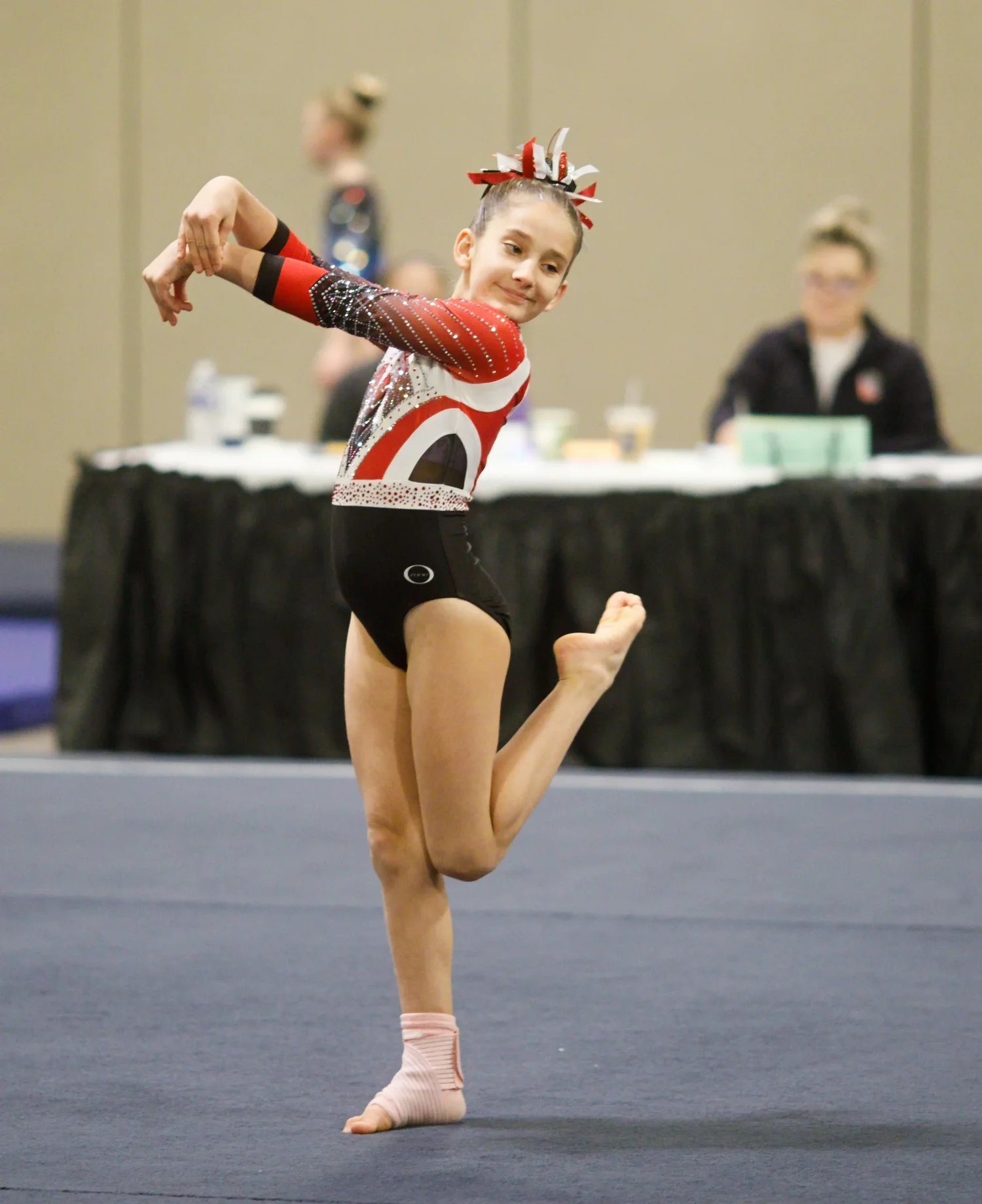 Gymnast in a red and silver leotard performing floor choreography with one leg raised.