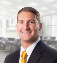 Tom wearing a dark striped suit with a white shirt and yellow tie smiling for the camera.