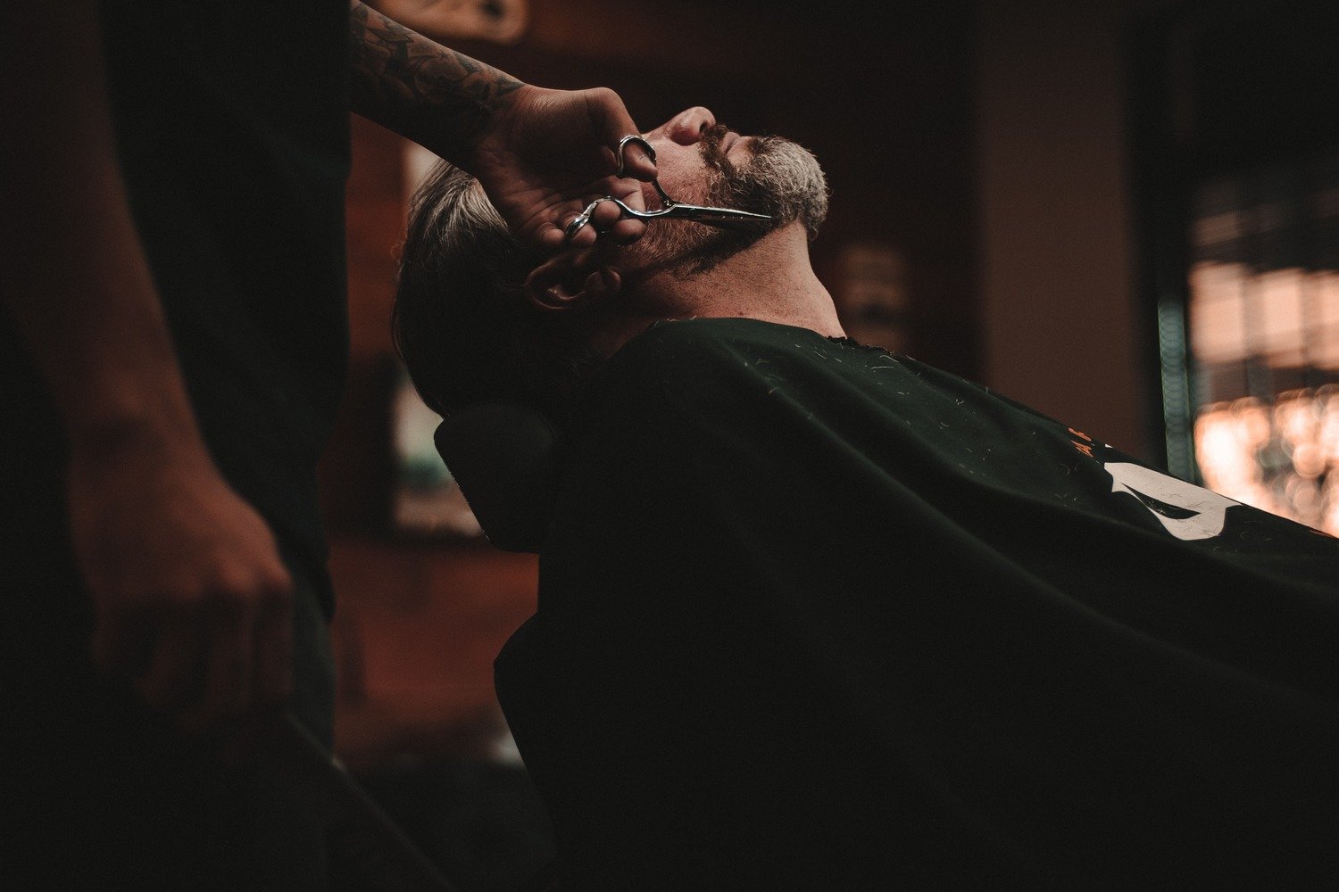 Barber trimming a client's beard with scissors in a barbershop.
