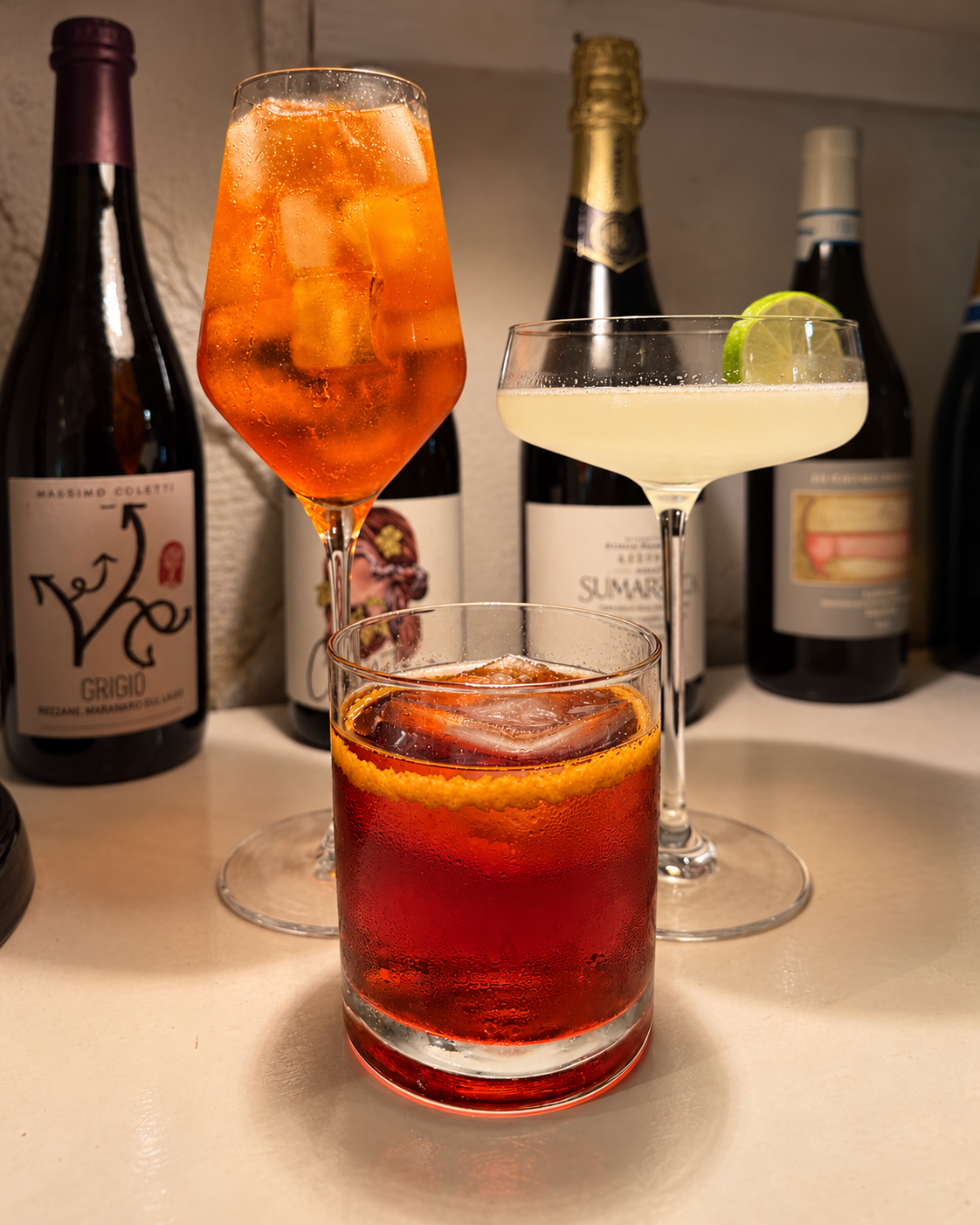 Three cocktails with citrus garnishes in front of Salut’s wine selection