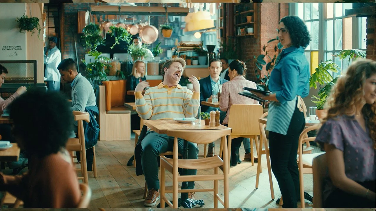 Actor Scott Watson crying and flailing at a restaurant table as Big Baby while waitress Sophia Blum looks on in the Plum Organics baby food commercial.